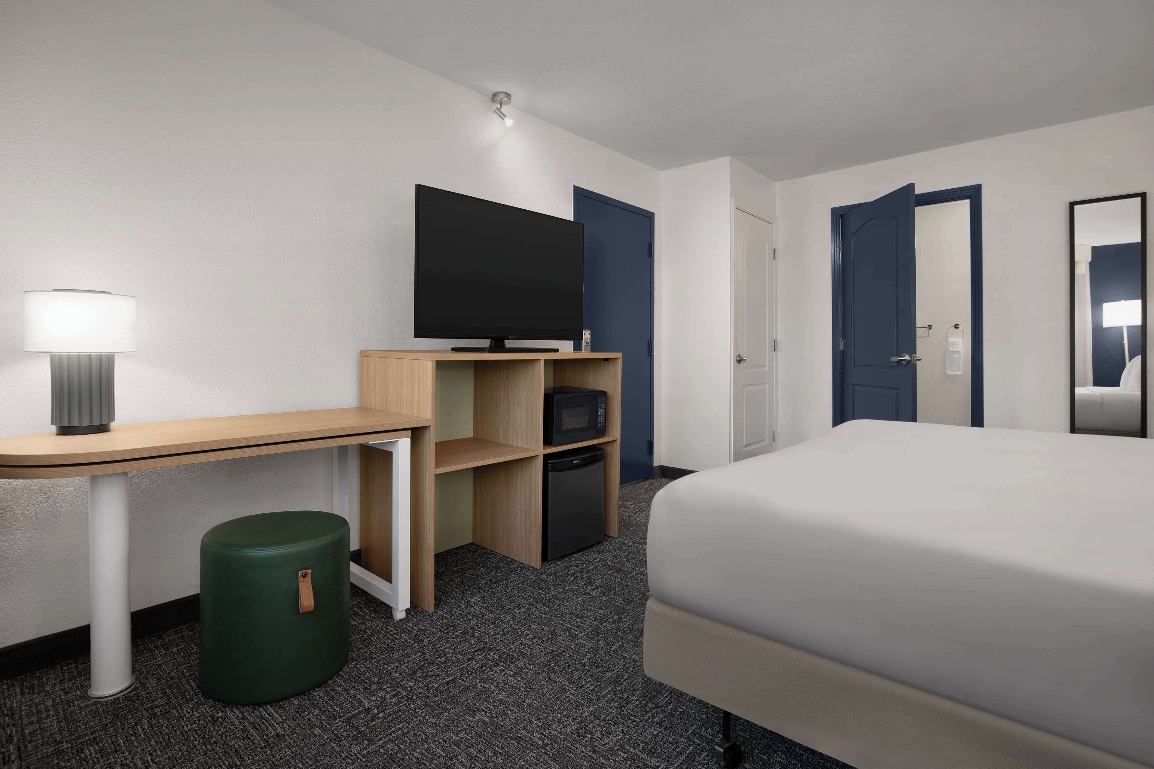 Een hotelkamer met een bed, een bureau met lamp, een televisie, een magnetron en een prullenbak.