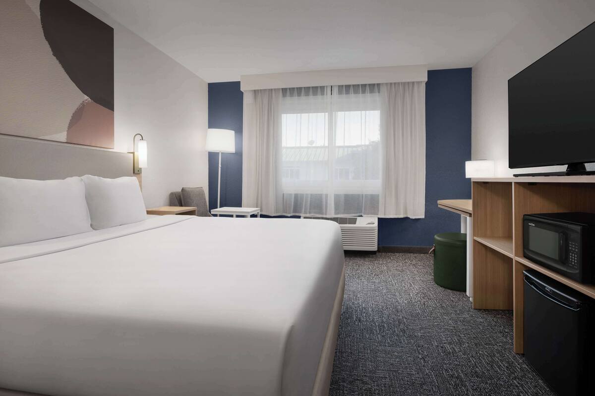 Ein Hotelzimmer mit einem großen Bett, einem Flachbildfernseher, einem Schreibtisch, einer Lampe und einem Fenster mit durchsichtigen Vorhängen.
