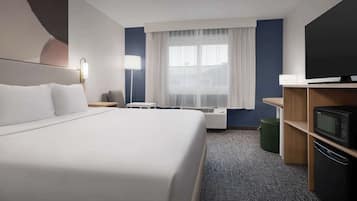 A hotel room with a large bed, a flat-screen TV, a desk, a lamp, and a window with sheer curtains.
