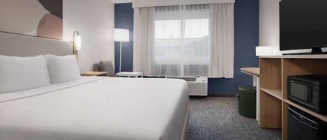 Quarto de hotel com uma cama grande, televisão de tela plana, escrivaninha, abajur e janela com cortinas leves.
