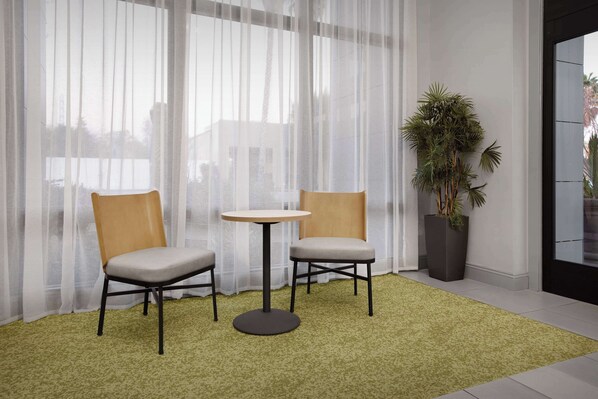 Una habitación con dos sillas, una mesita pequeña, cortinas de gasa, una alfombra verde y una planta en maceta.