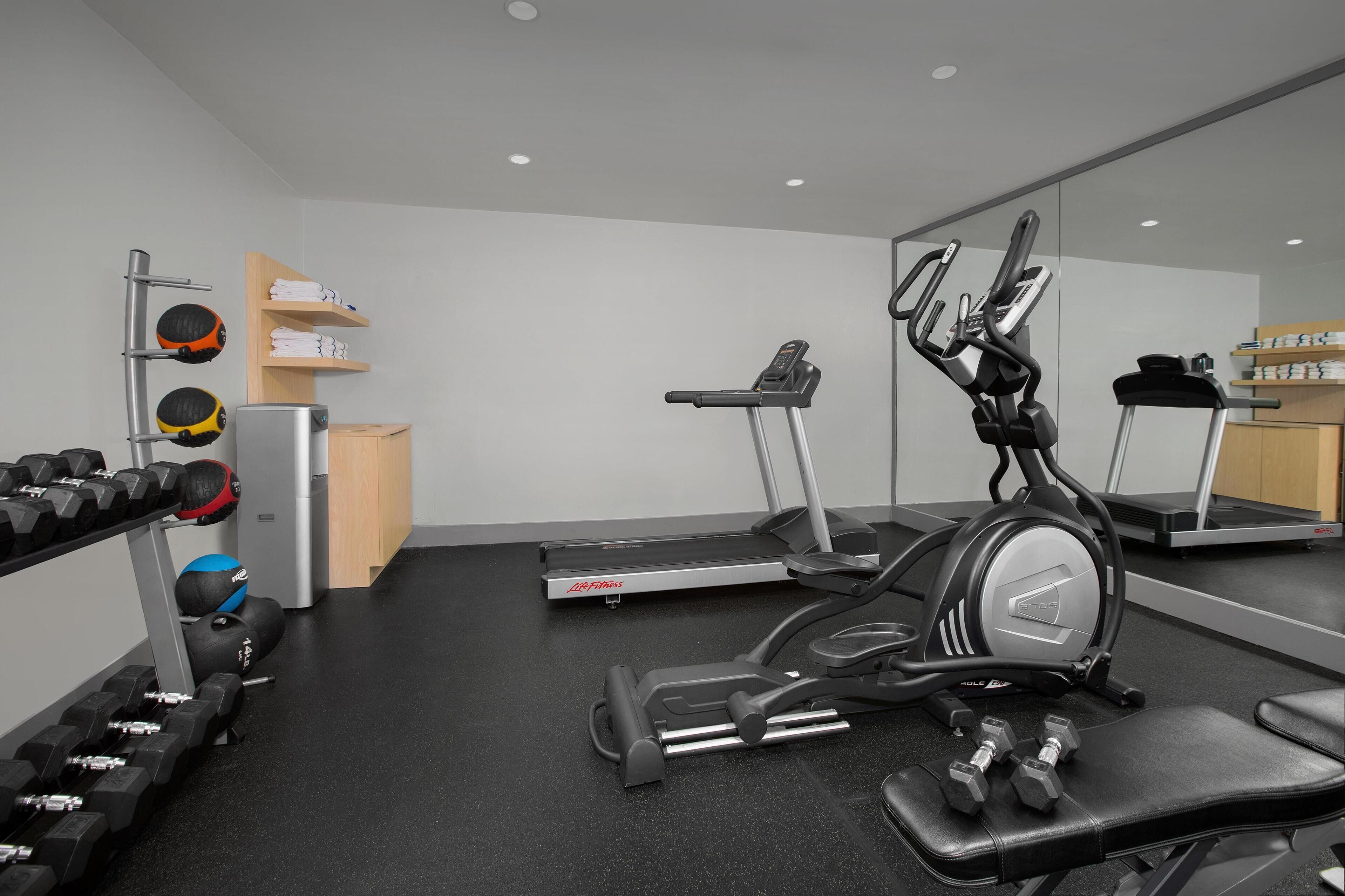 A modern gym with cardio equipment, dumbbells, and a weight rack.