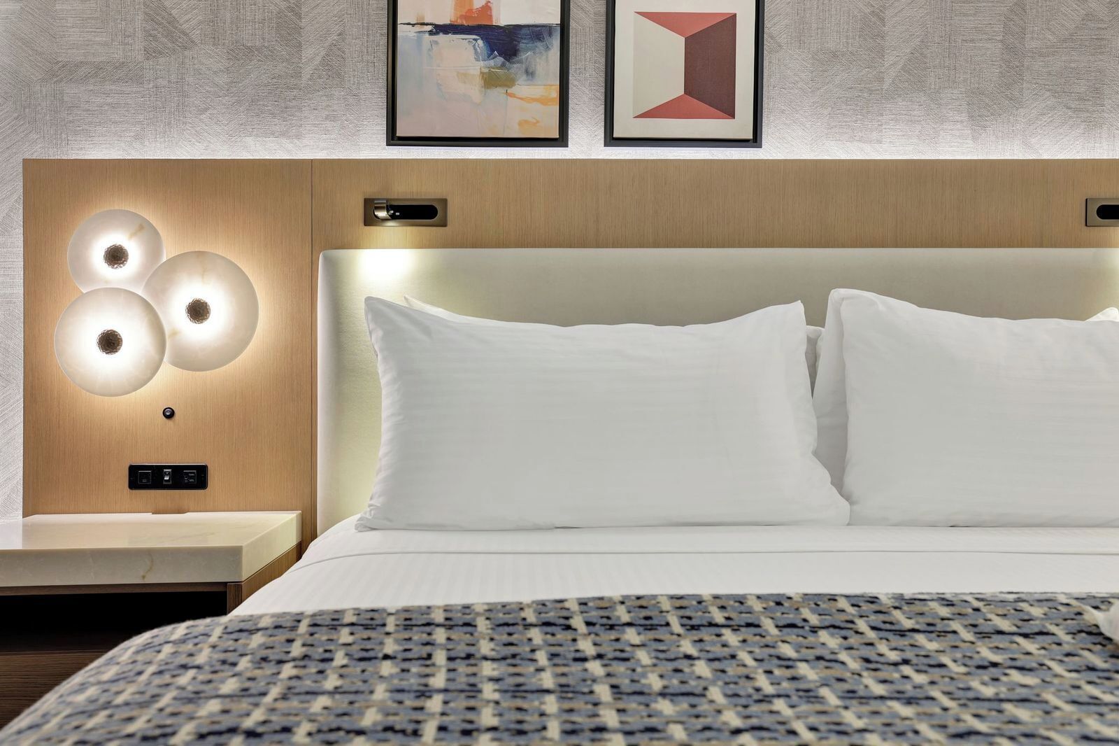 A hotel room with a bed, a nightstand, a wall-mounted light fixture, and two framed artworks.