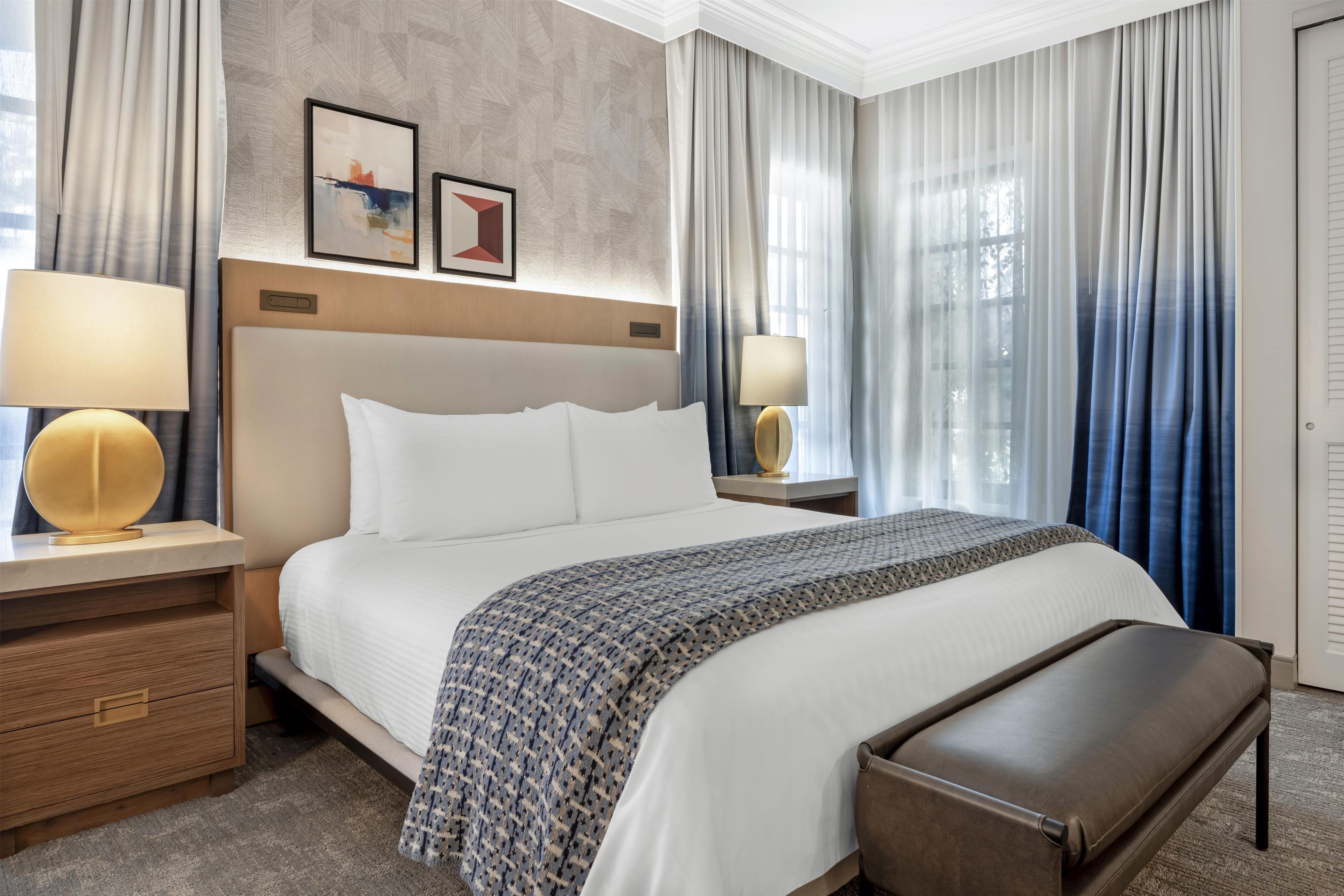 A hotel room with a large bed, two bedside tables with lamps, a wooden bench, a patterned throw blanket, and a view of the outdoors through sheer curtains.