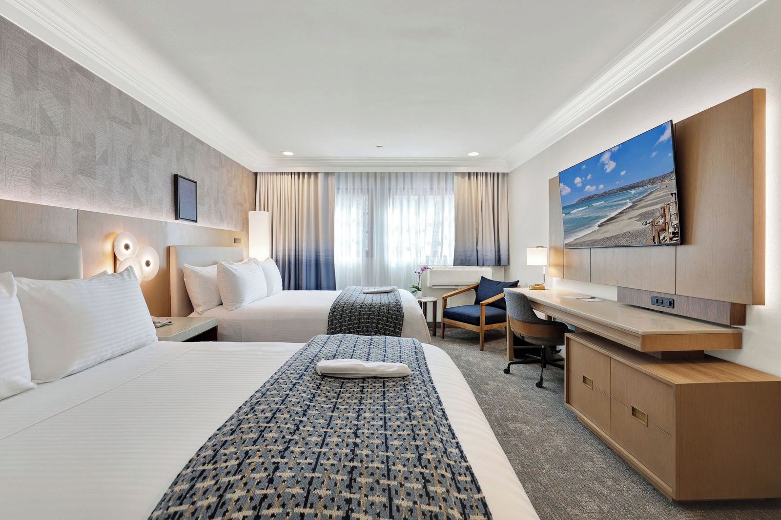 Une chambre d’hôtel avec deux lits, un bureau, une chaise, une télévision et un tableau représentant une plage.