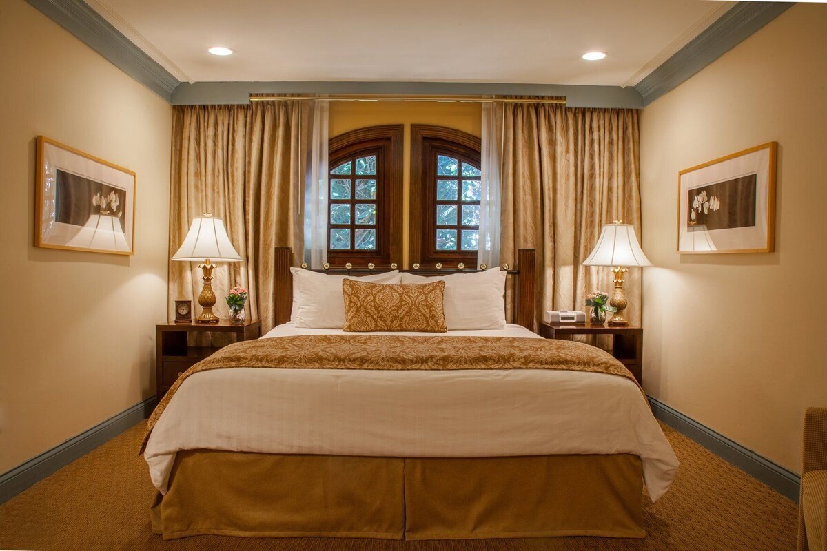 Un lit double avec une couvre-lit à motifs, deux tables de chevet avec des lampes, une fenêtre avec des rideaux et des tableaux encadrés accrochés aux murs.