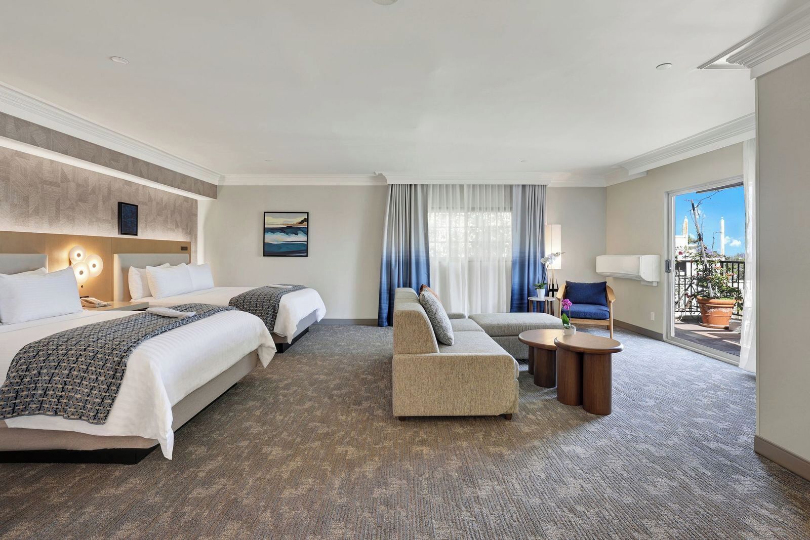 A hotel room with two beds, a sofa, a small table, and a balcony with potted plants.