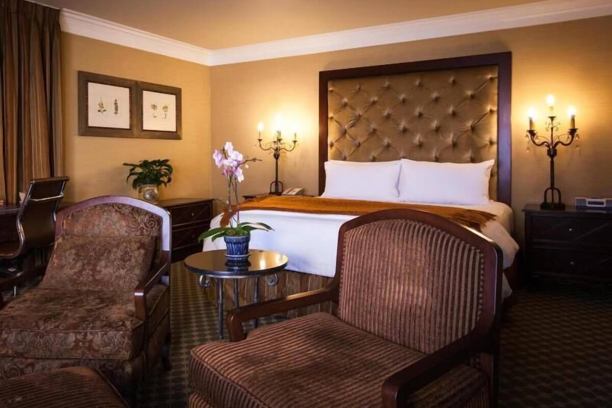 Une chambre d’hôtel avec un grand lit, deux chaises, une petite table avec un vase et des tableaux encadrés au mur.