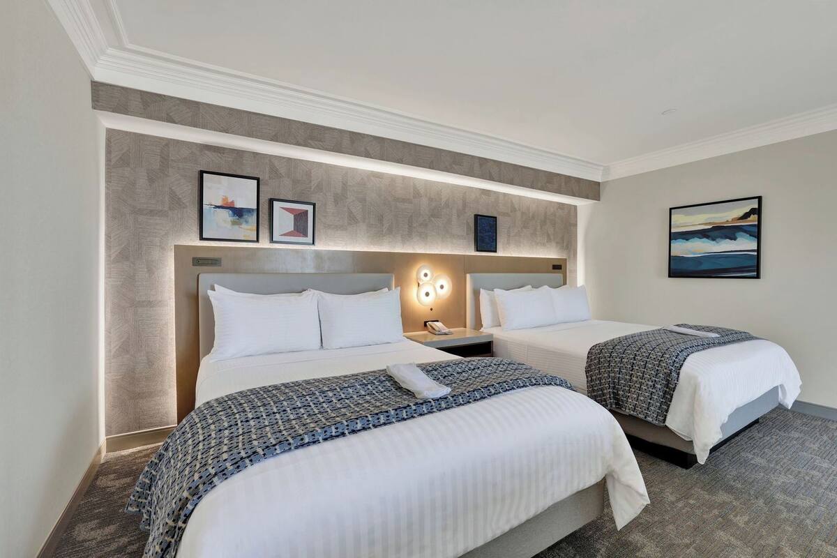 Une chambre d’hôtel avec deux lits, une tête de lit dotée d’un éclairage intégré, une petite table de chevet avec un téléphone et une œuvre d’art accrochée au mur.