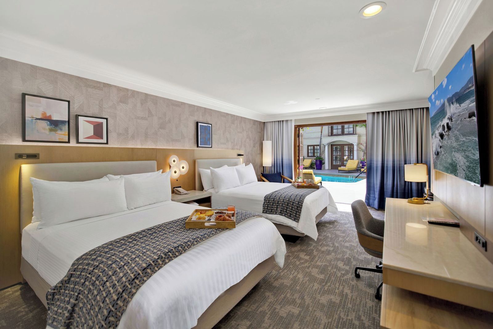 A hotel room with two beds, a desk, a chair, a TV, and a view of a pool.