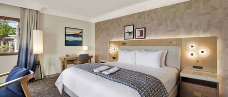 Ein Hotelzimmer mit einem großen Bett, Nachttischen, einem Schreibtisch, einem Sessel und Blick ins Freie.