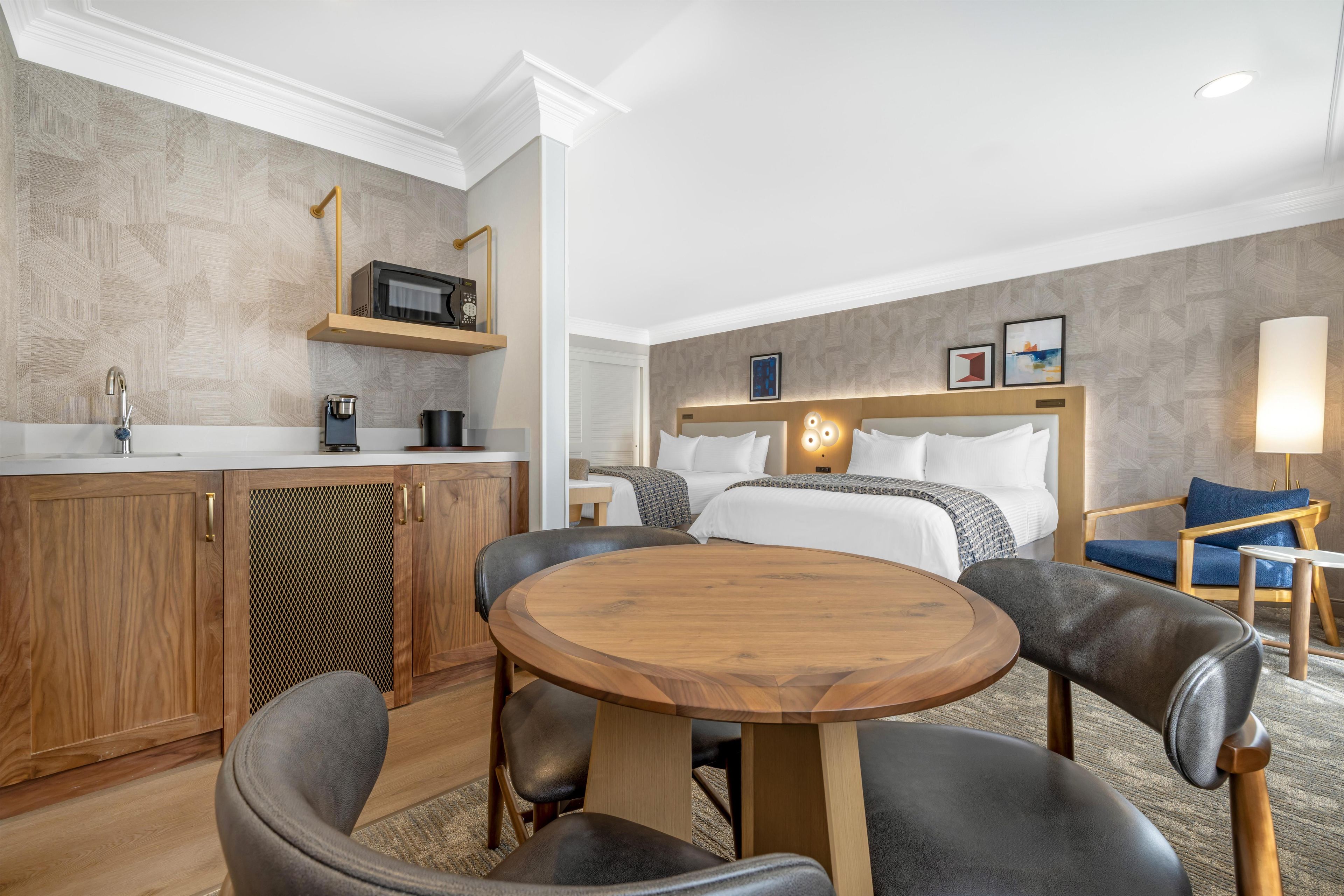 A hotel room with a round wooden table, two chairs, a bed with white bedding, a bedside table with a lamp, a small shelf with a microwave and coffee maker, and a wall-mounted TV.