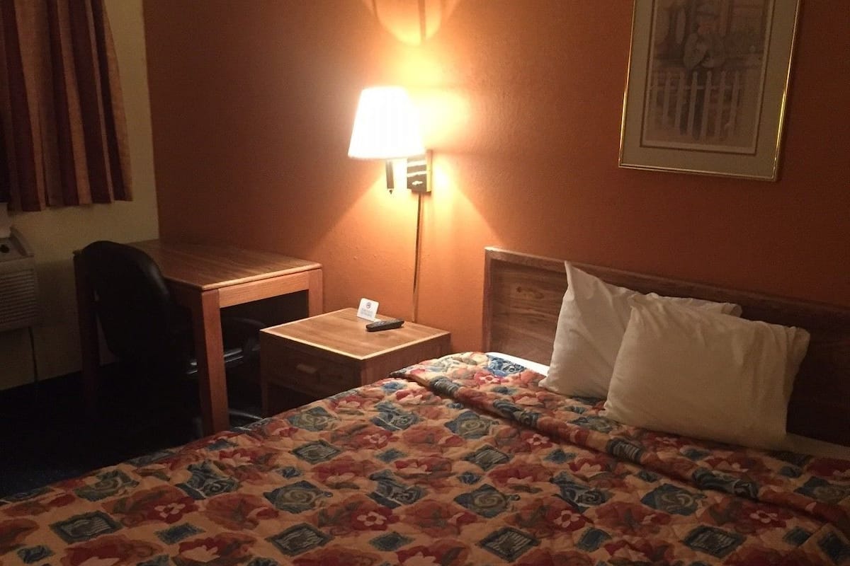 A hotel room with a bed, a desk, a chair, a nightstand, a lamp, and a framed picture on the wall.