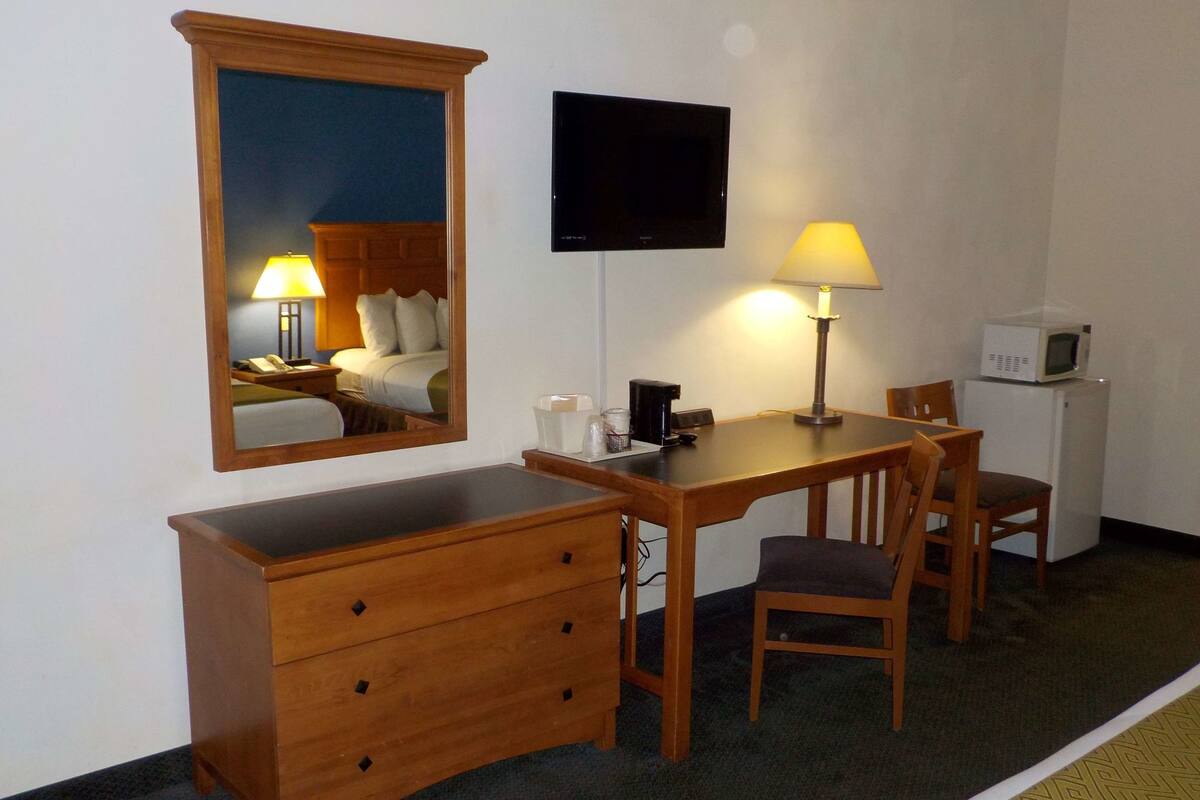 A hotel room with a wooden desk, two chairs, a television, a mirror, a lamp, a microwave, and a small refrigerator.