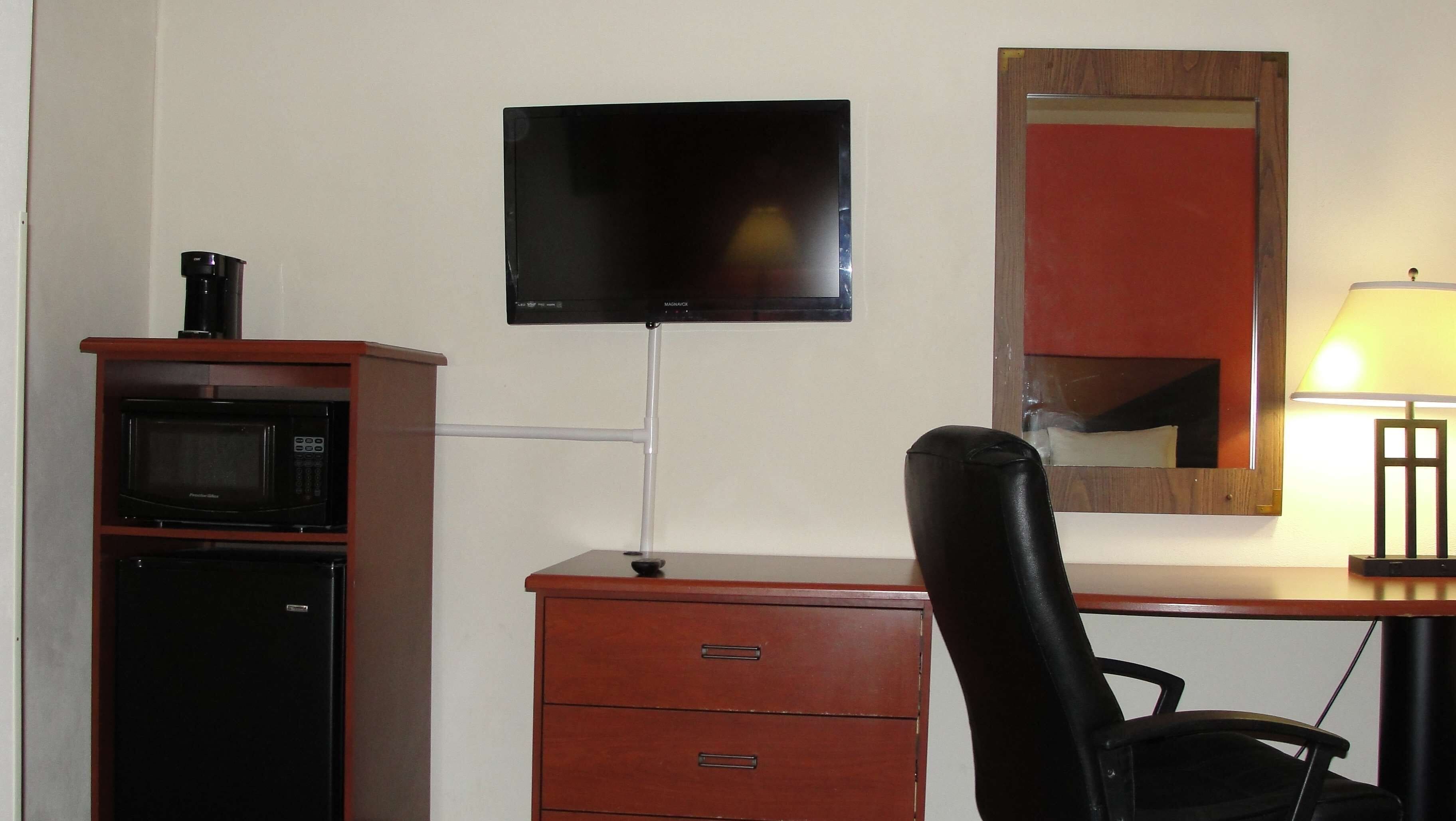A hotel room with a wooden desk, a television mounted on the wall, a microwave, a chair, and a lamp.