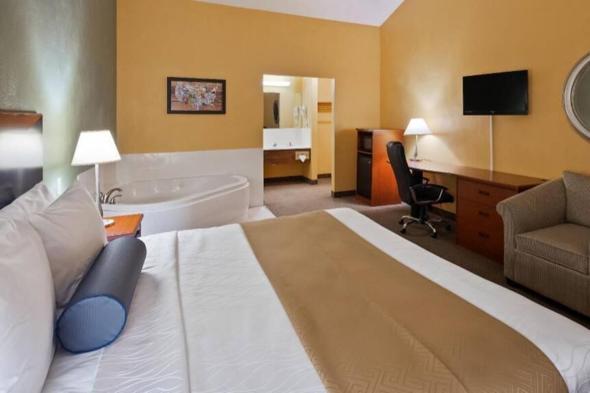 A hotel room with a large bed, a desk, a chair, a TV, a mirror, and a bathroom with a bathtub and sink.