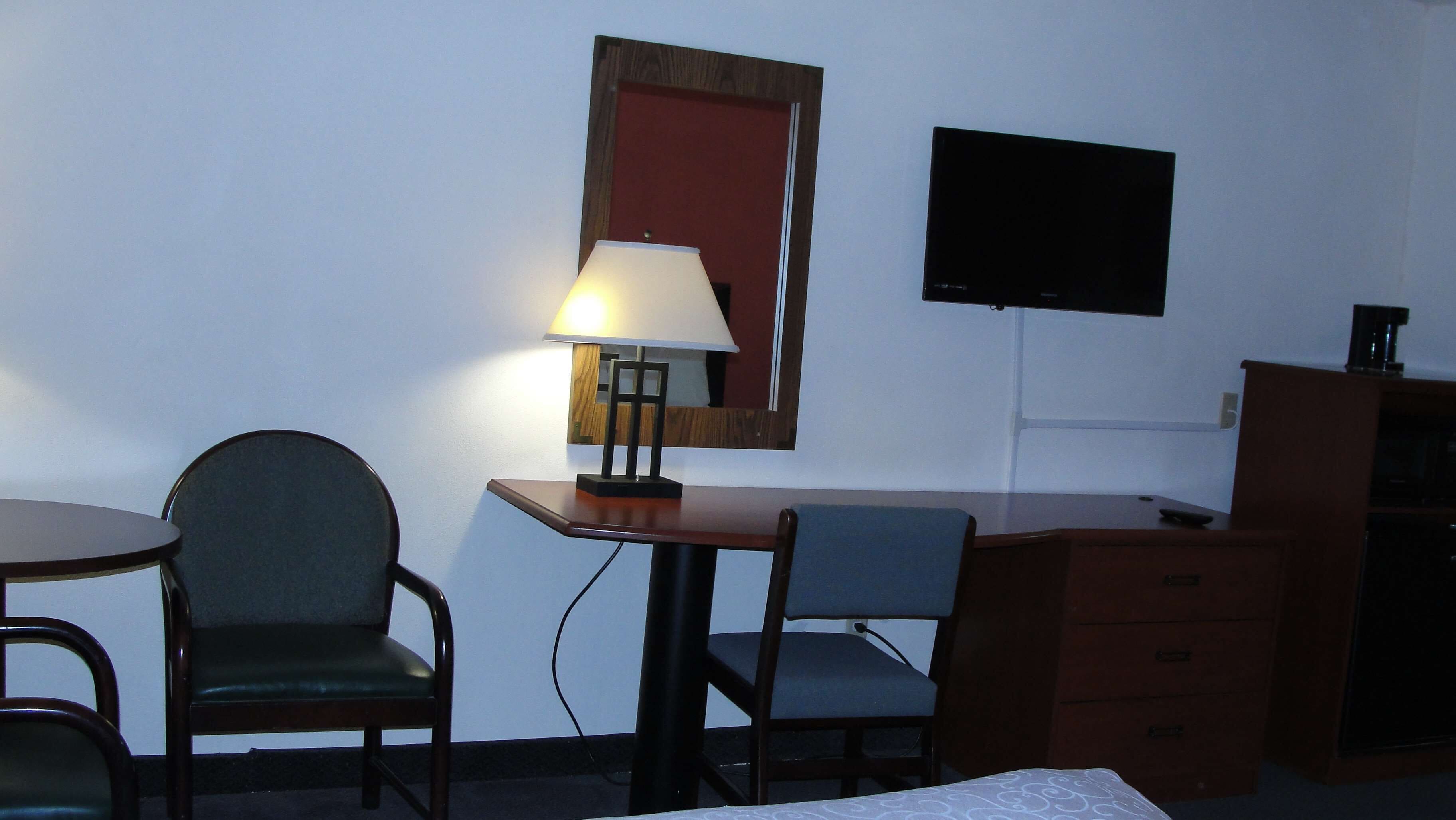 A small, simple room with a desk, two chairs, a lamp, a TV, and a dresser.