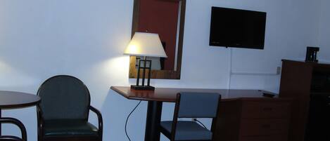 A small, simple room with a desk, two chairs, a lamp, a TV, and a dresser.