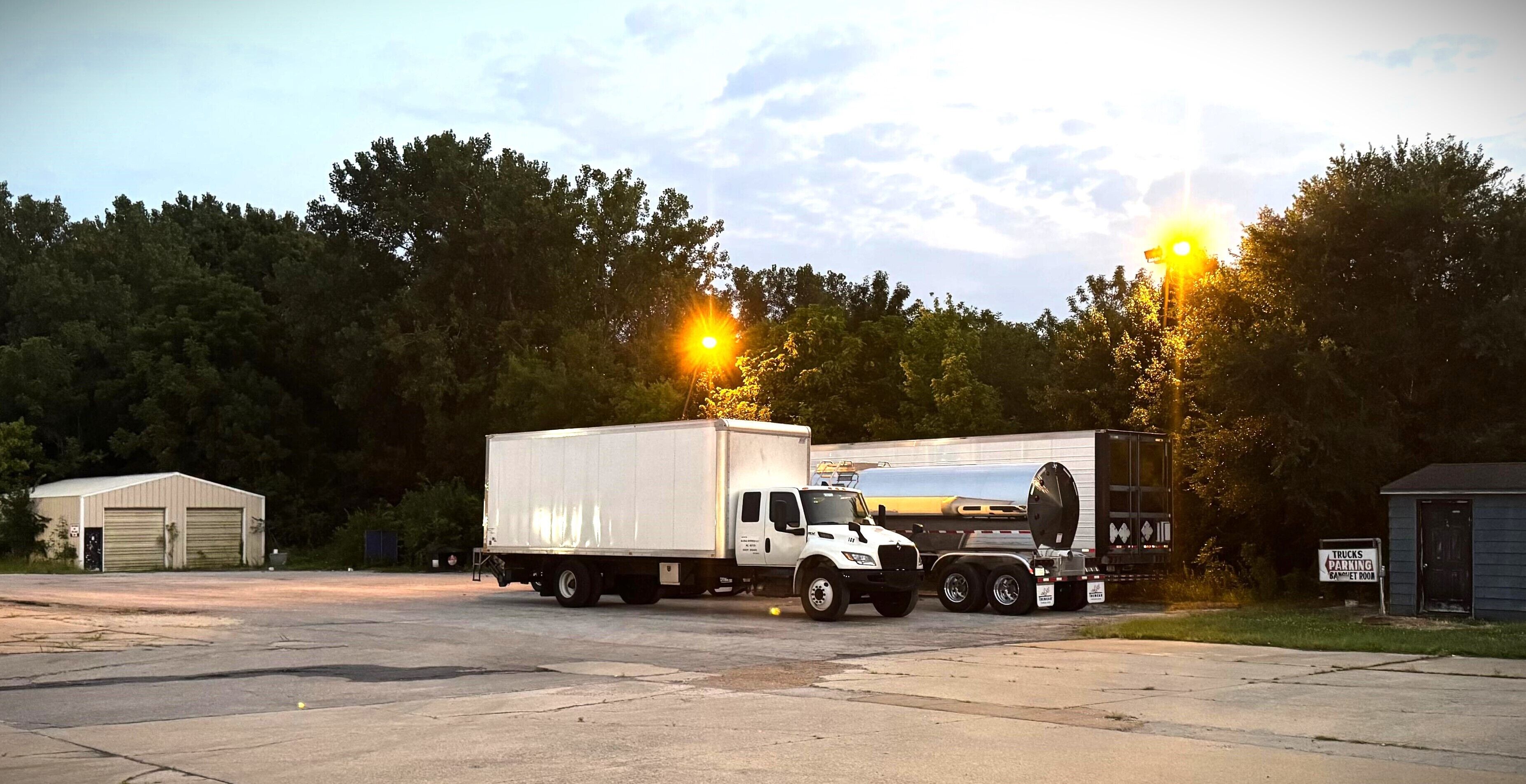 A parking lot with a white truck and a smaller vehicle, surrounded by trees and a building with a sign that reads 'TRUCKS PARKING'.