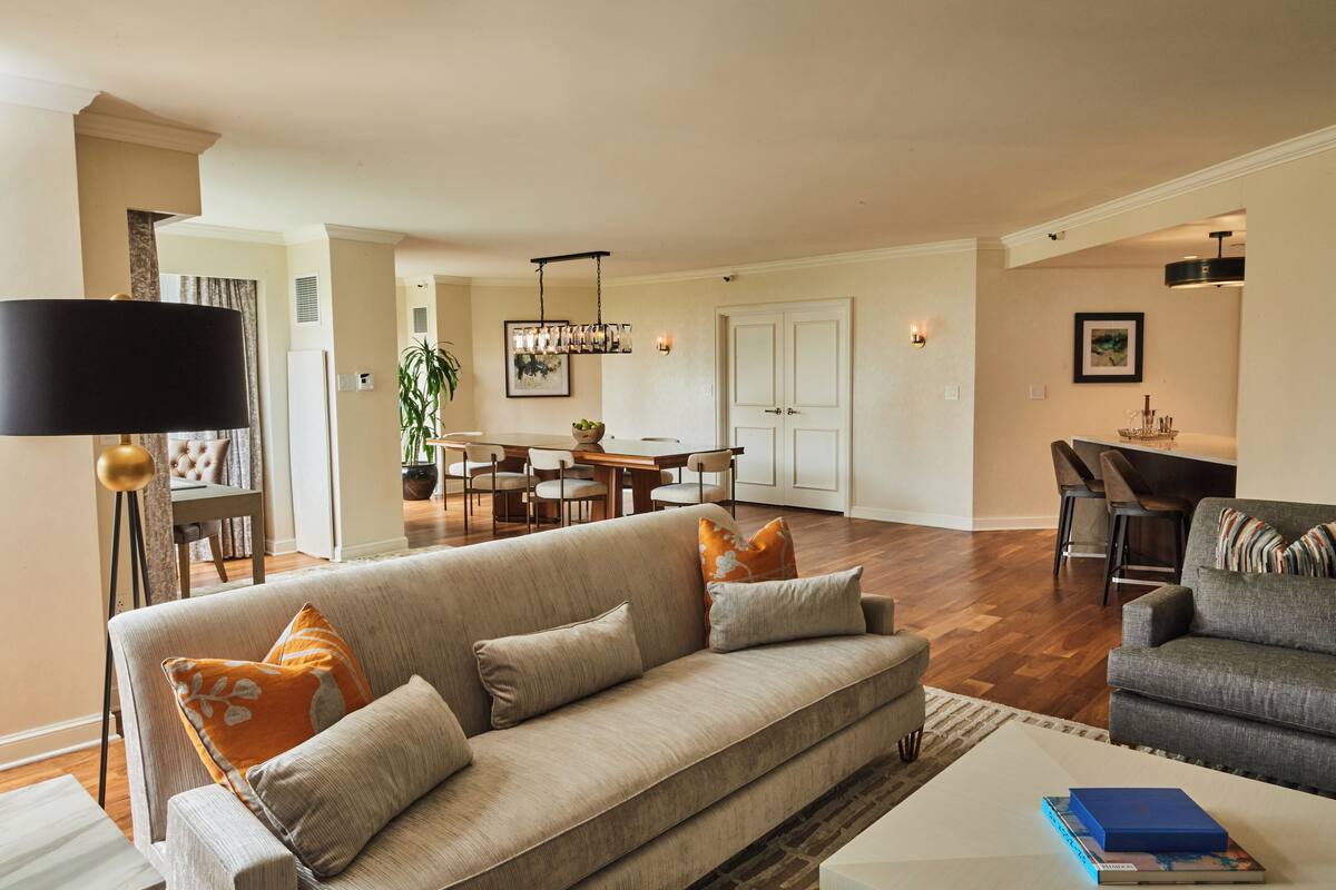 A spacious living room with a sofa, armchairs, and a coffee table. There is a dining area with a table and chairs, and a kitchen in the background.
