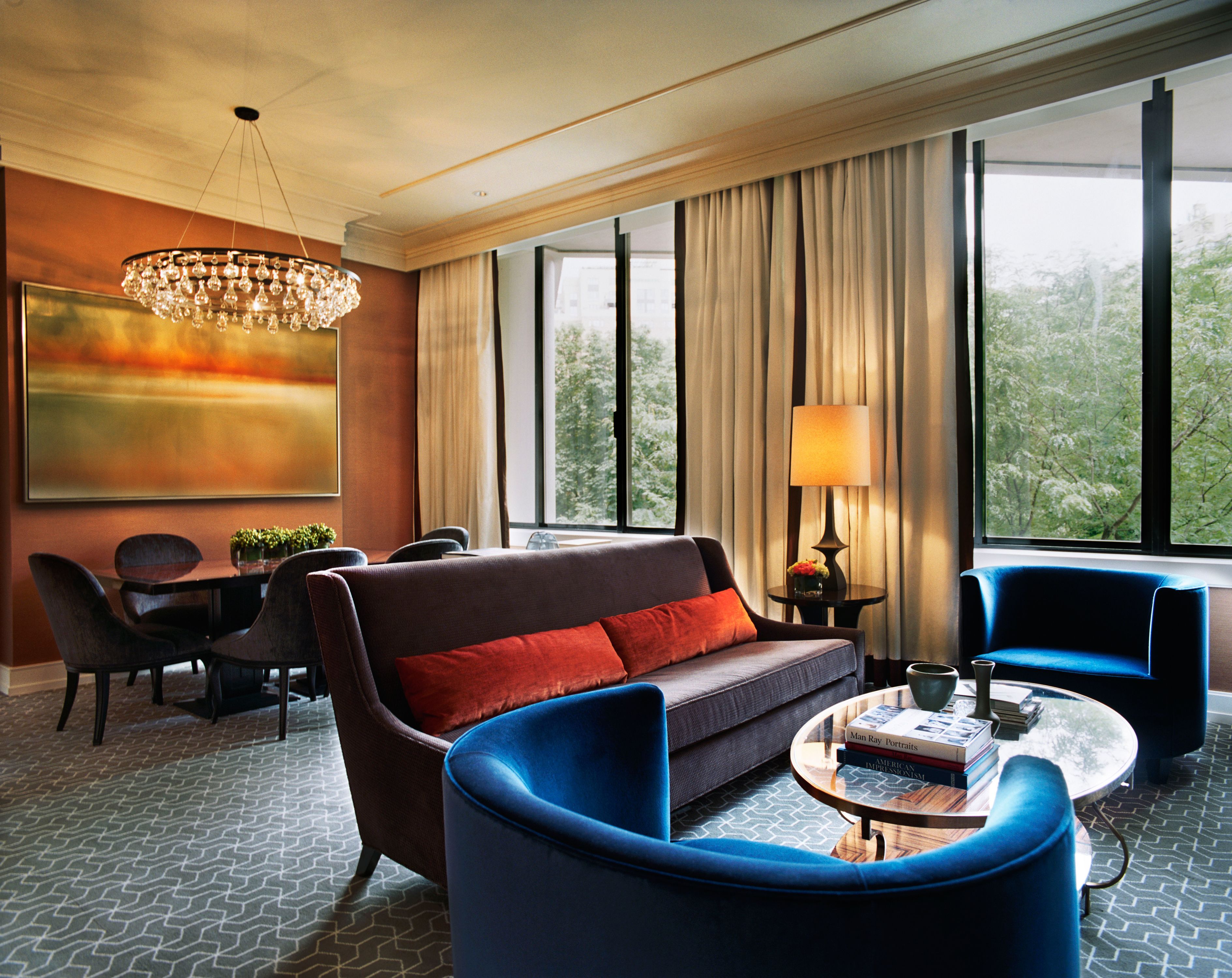 A modern hotel room with a sofa, two armchairs, a coffee table, a dining area with chairs, a chandelier, and large windows with curtains.