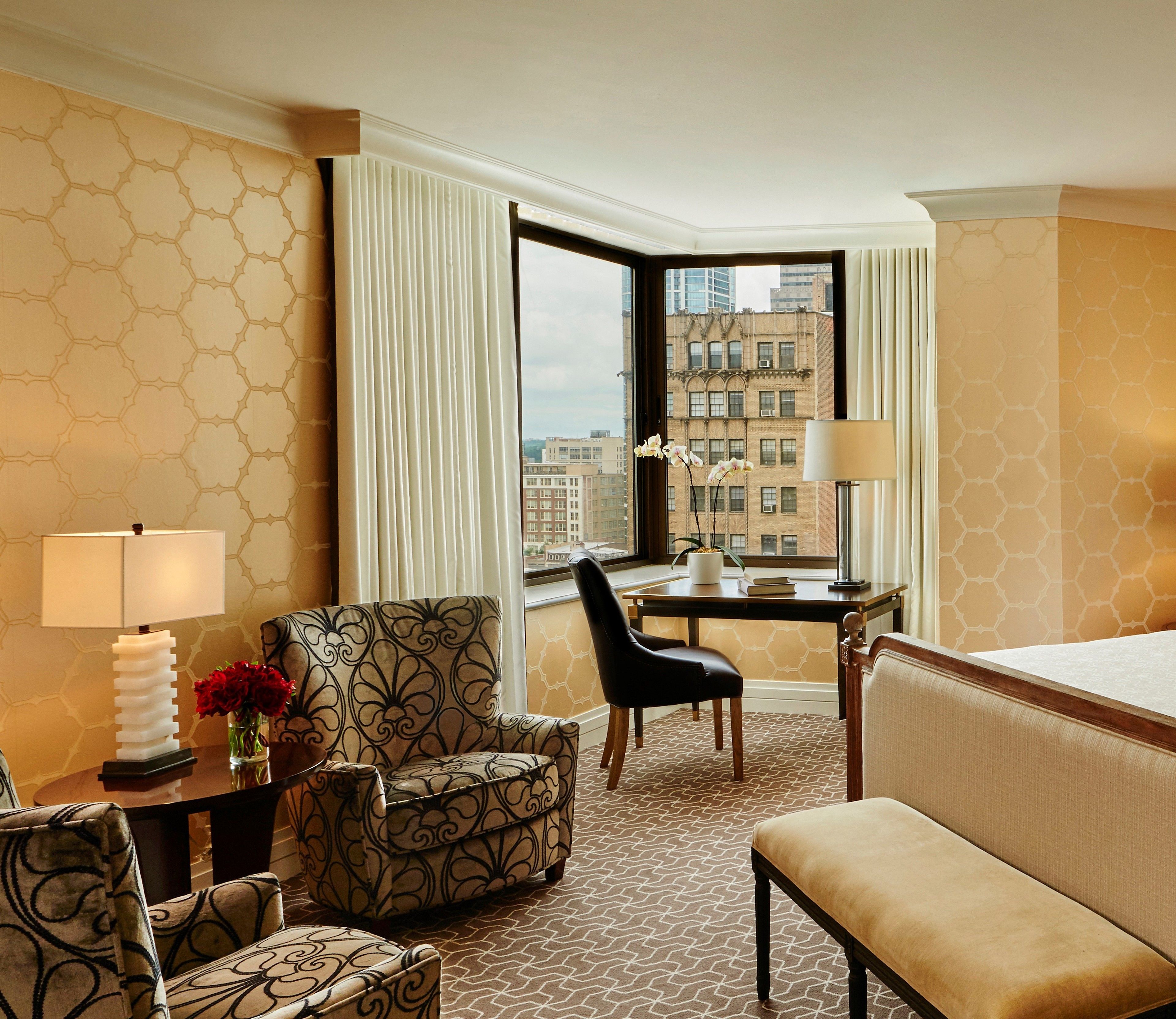 A hotel room with a corner bed, a desk with a lamp, a chair, a sofa, and a view of buildings through a window.