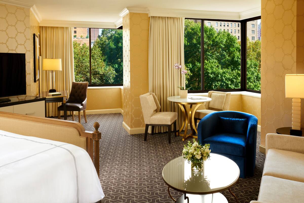 A hotel room with a large bed, two armchairs, a sofa, a small round table, a TV, and a window with a view of trees and buildings.