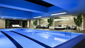 A modern indoor swimming pool with a blue floor, surrounded by seating areas and potted plants.