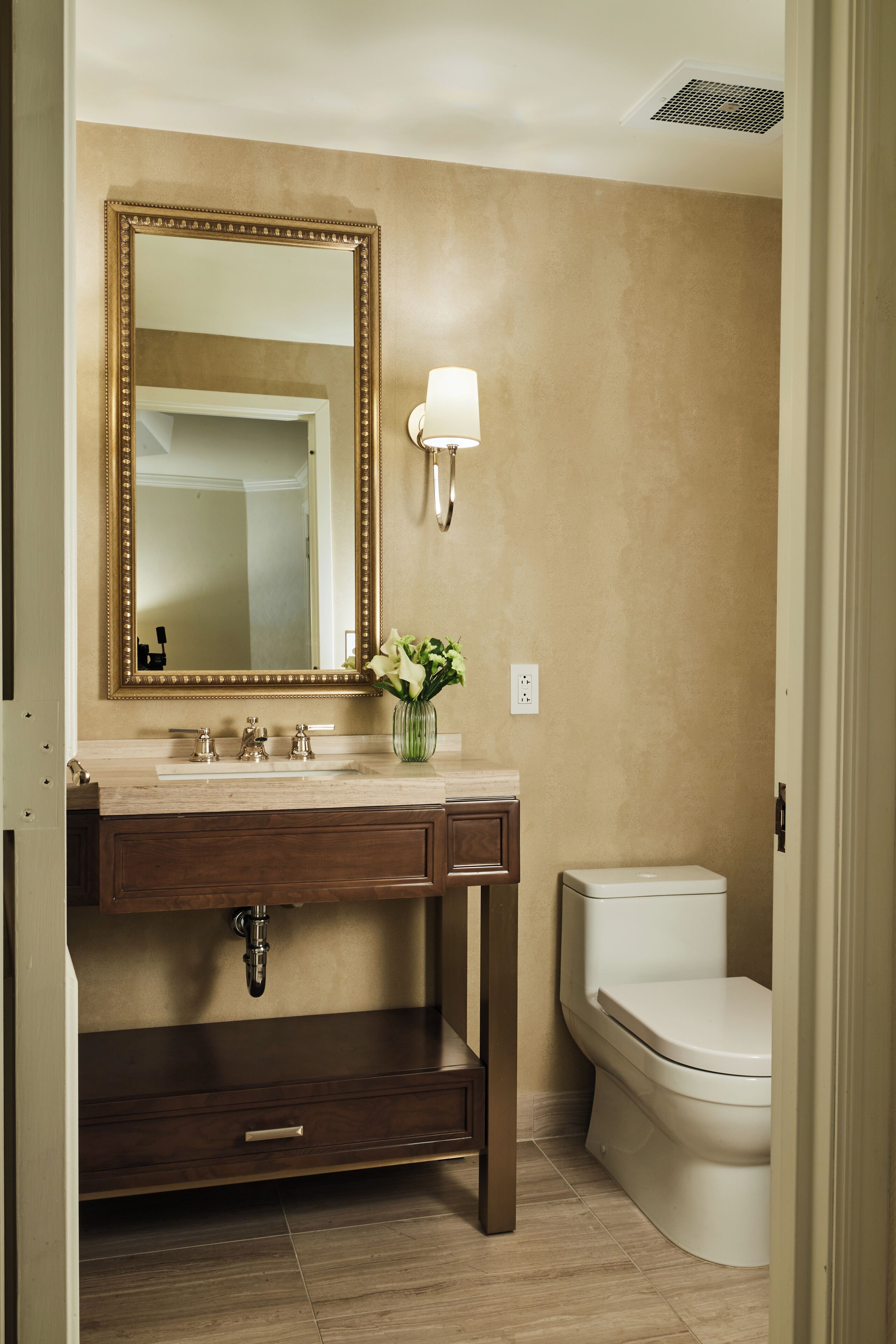 Un baño con pared color beige, un espejo grande, un lavamanos con un florero y un inodoro, además de un mueble de tocador de madera.
