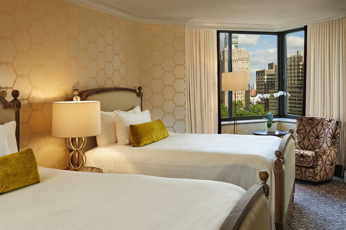 A hotel room with two beds, a large window with a city view, a patterned armchair, and a lamp on a bedside table.