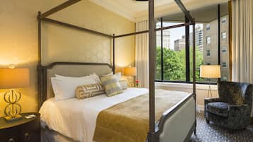 A four-poster bed with a canopy, a nightstand with a lamp, a chair, and a view of the city through large windows.