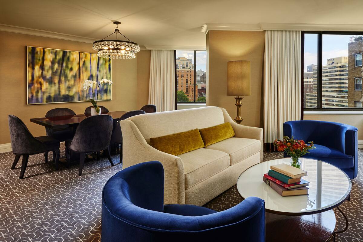 A hotel room with a dining area, a sofa, two armchairs, a coffee table with books, a chandelier, a large window with a city view, and a painting on the wall.