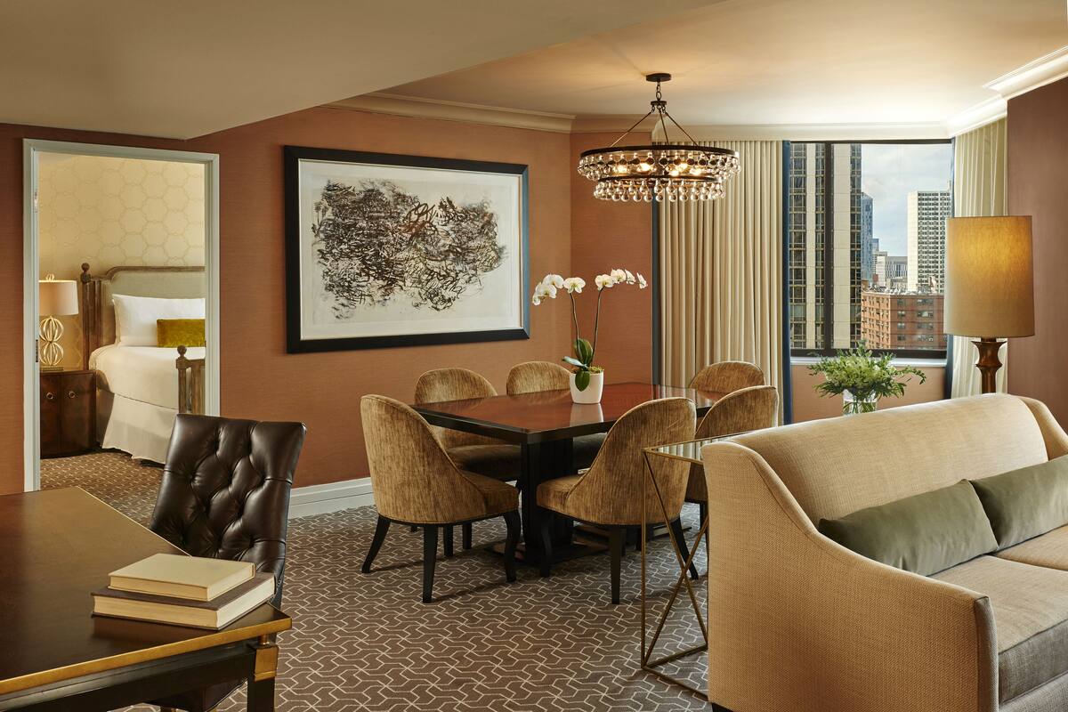 A hotel room with a dining table, chairs, a sofa, a chandelier, and a large window with a city view.