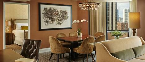 A hotel room with a dining table, chairs, a sofa, a chandelier, and a large window with a city view.