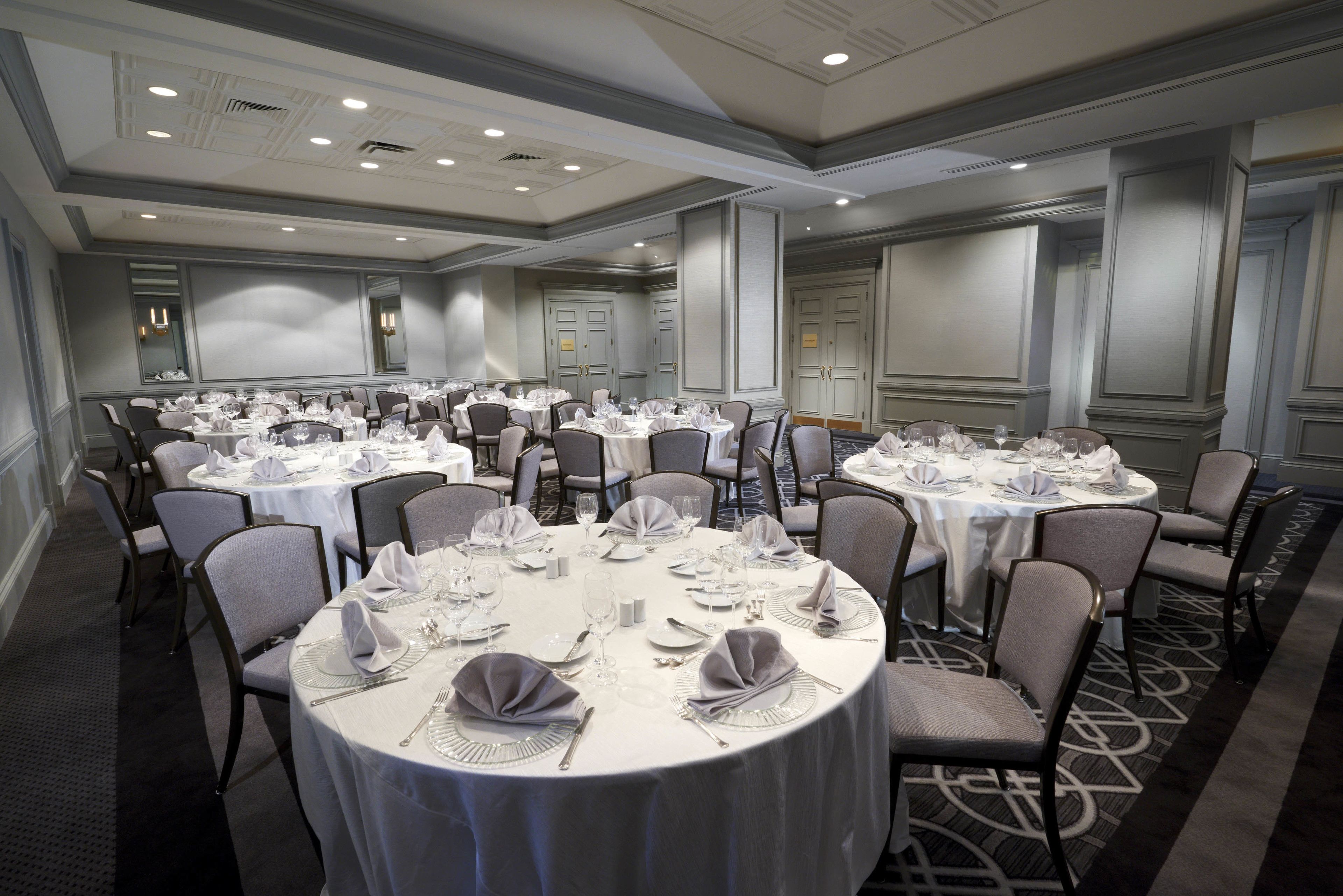 Une salle de conférence dotée de plusieurs tables rondes, chacune recouverte d’une nappe blanche et agrémentée de chaises et du nécessaire pour le service à table.