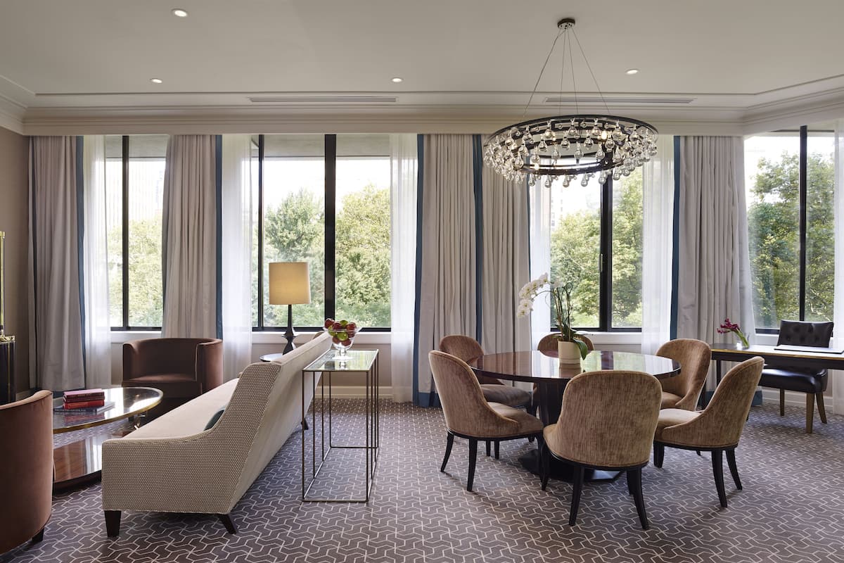 A hotel room with a large window, a chandelier, a sofa, a dining table with chairs, and a TV.