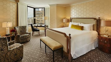 Ein Hotelzimmer mit einem großen Bett, einem Schreibtisch, zwei Sesseln, einer Bank und Blick auf die Stadt durch ein großes Fenster.