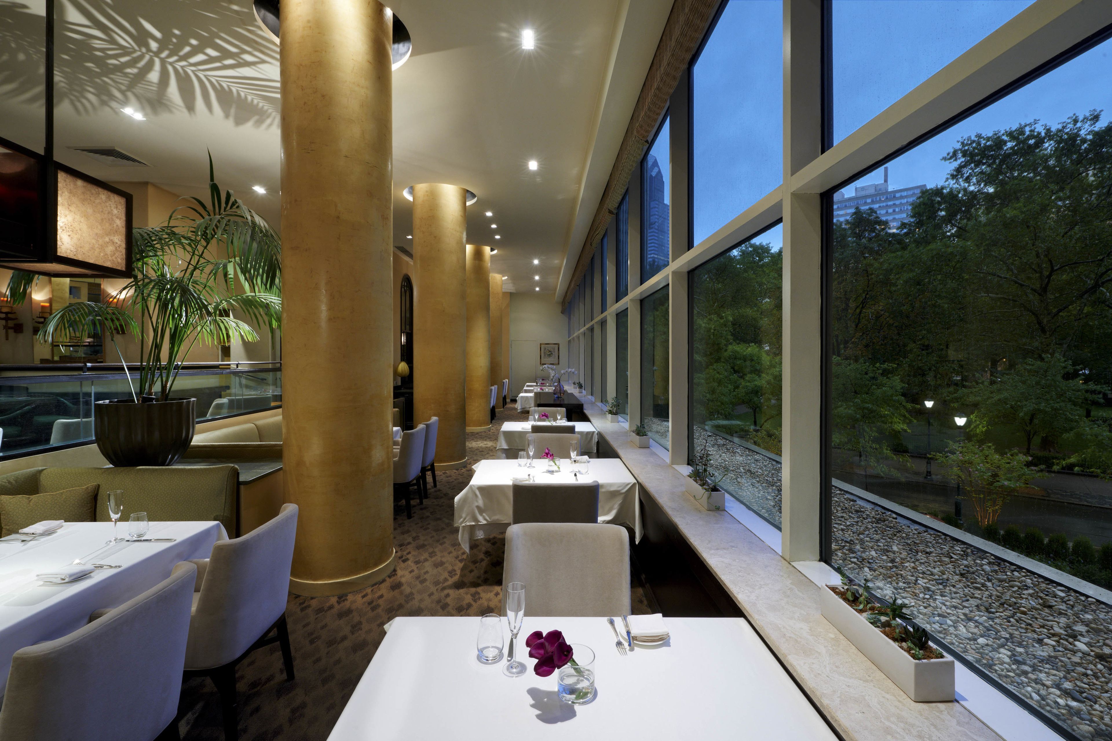 A restaurant with white tables, beige chairs, and large windows overlooking a garden.