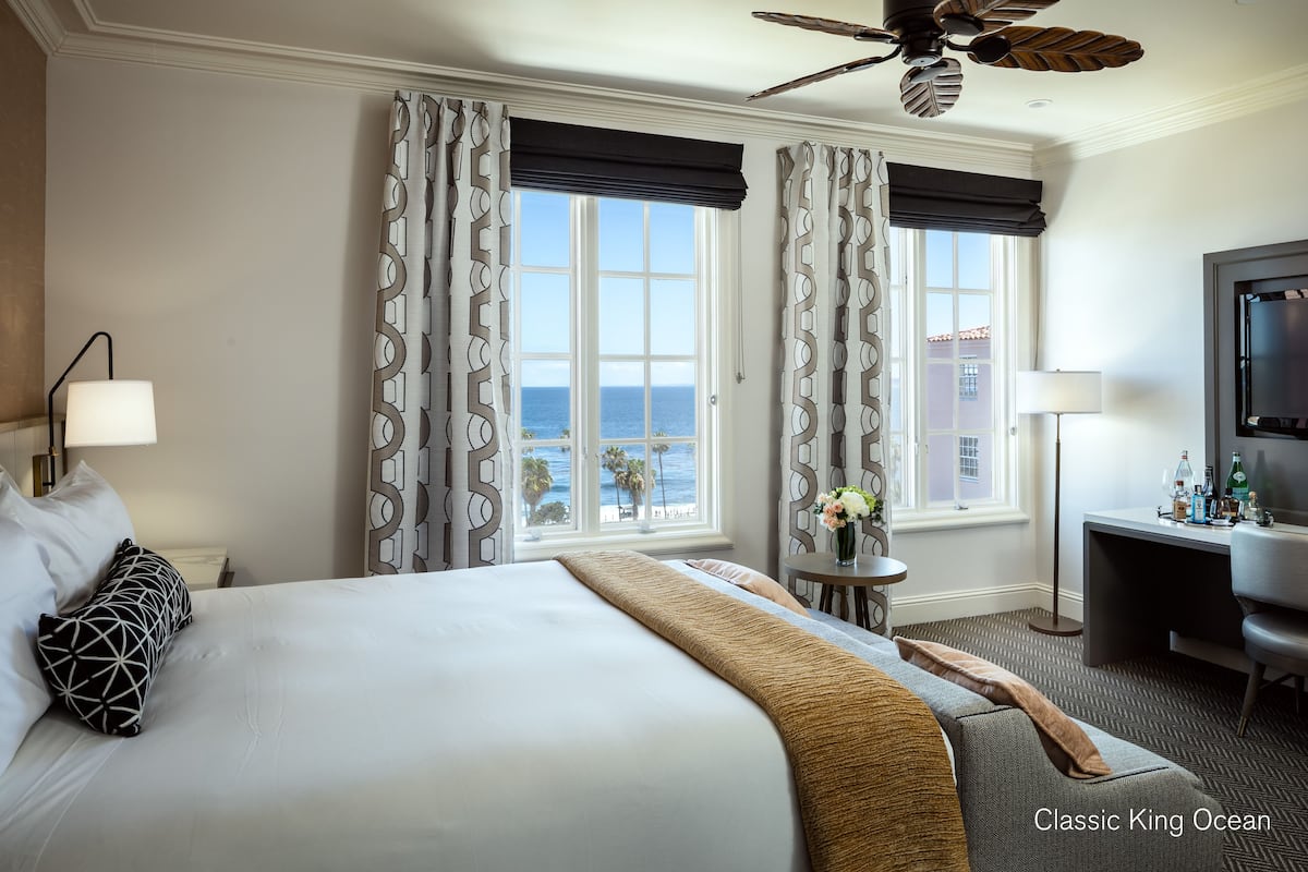 A hotel room with a large bed, two windows with ocean views, a desk with a TV, and a ceiling fan.