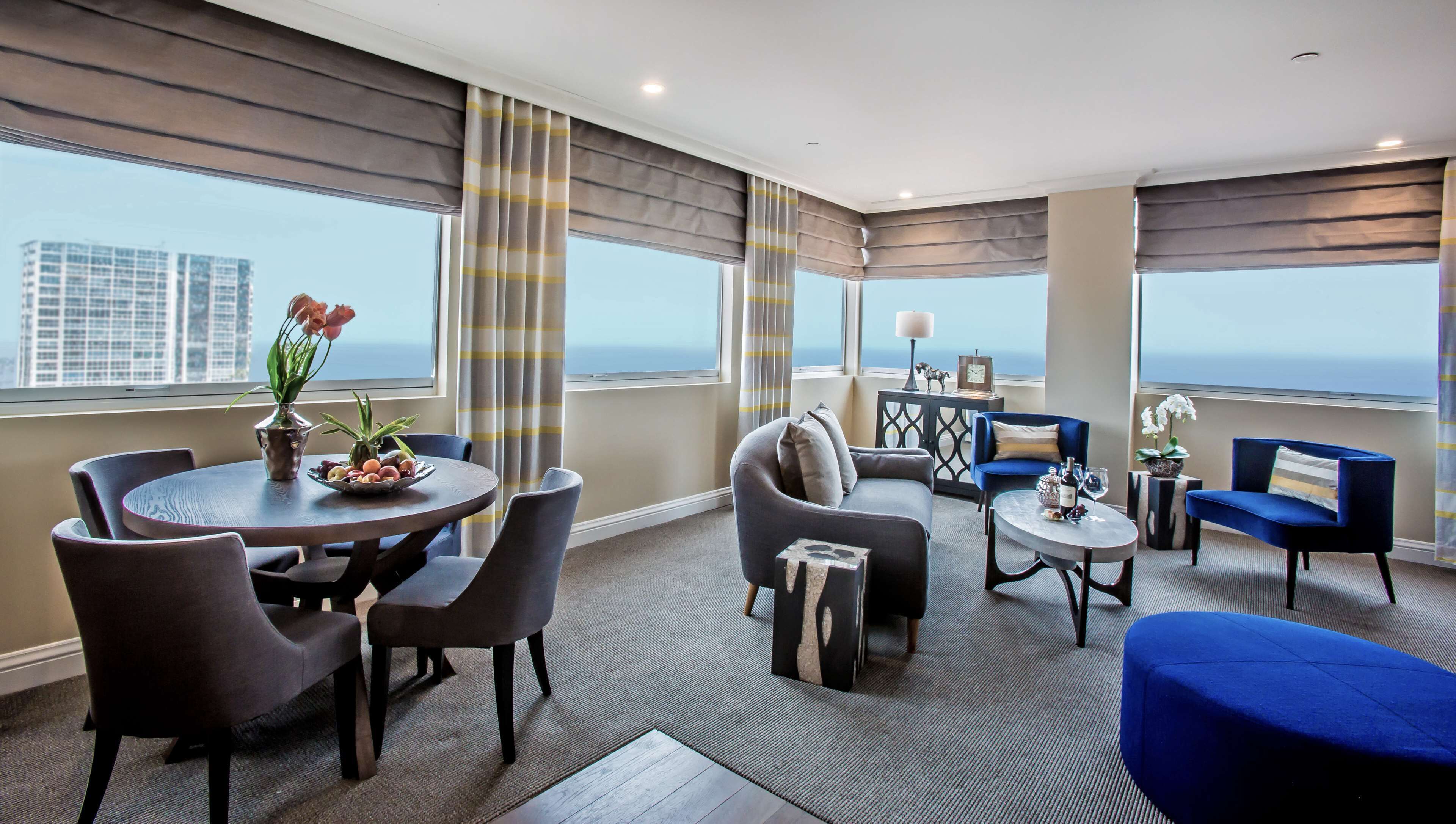 A modern hotel room with a dining area, seating, and large windows offering a city view.