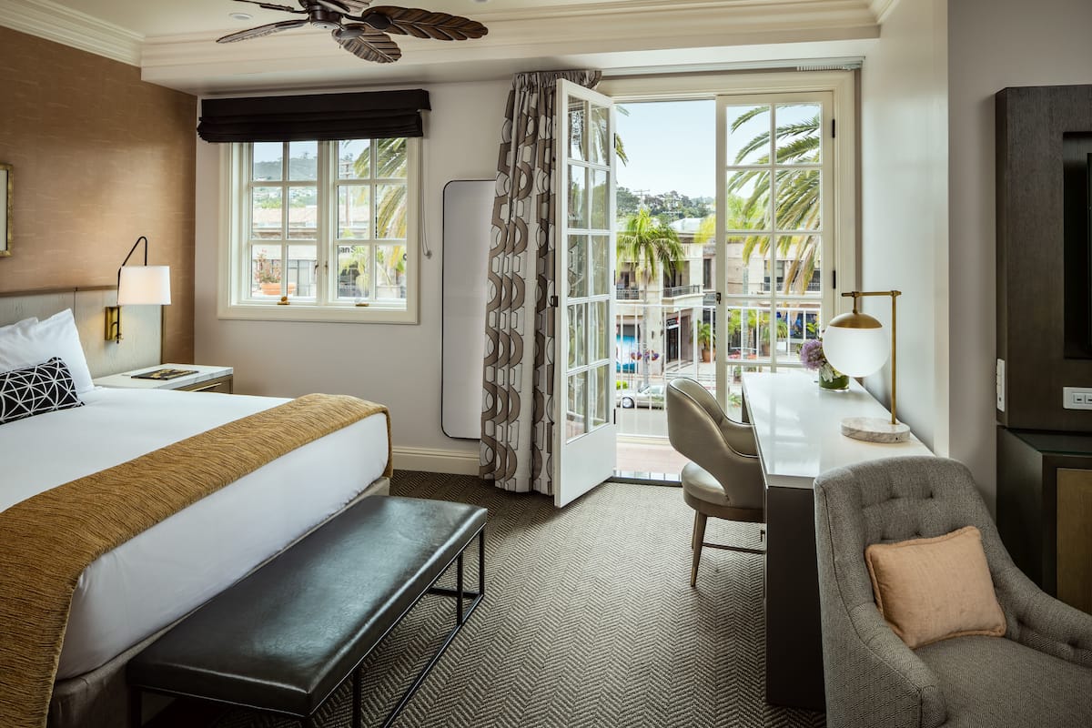 A hotel room with a bed, bench, desk, chair, and a view of the outside.