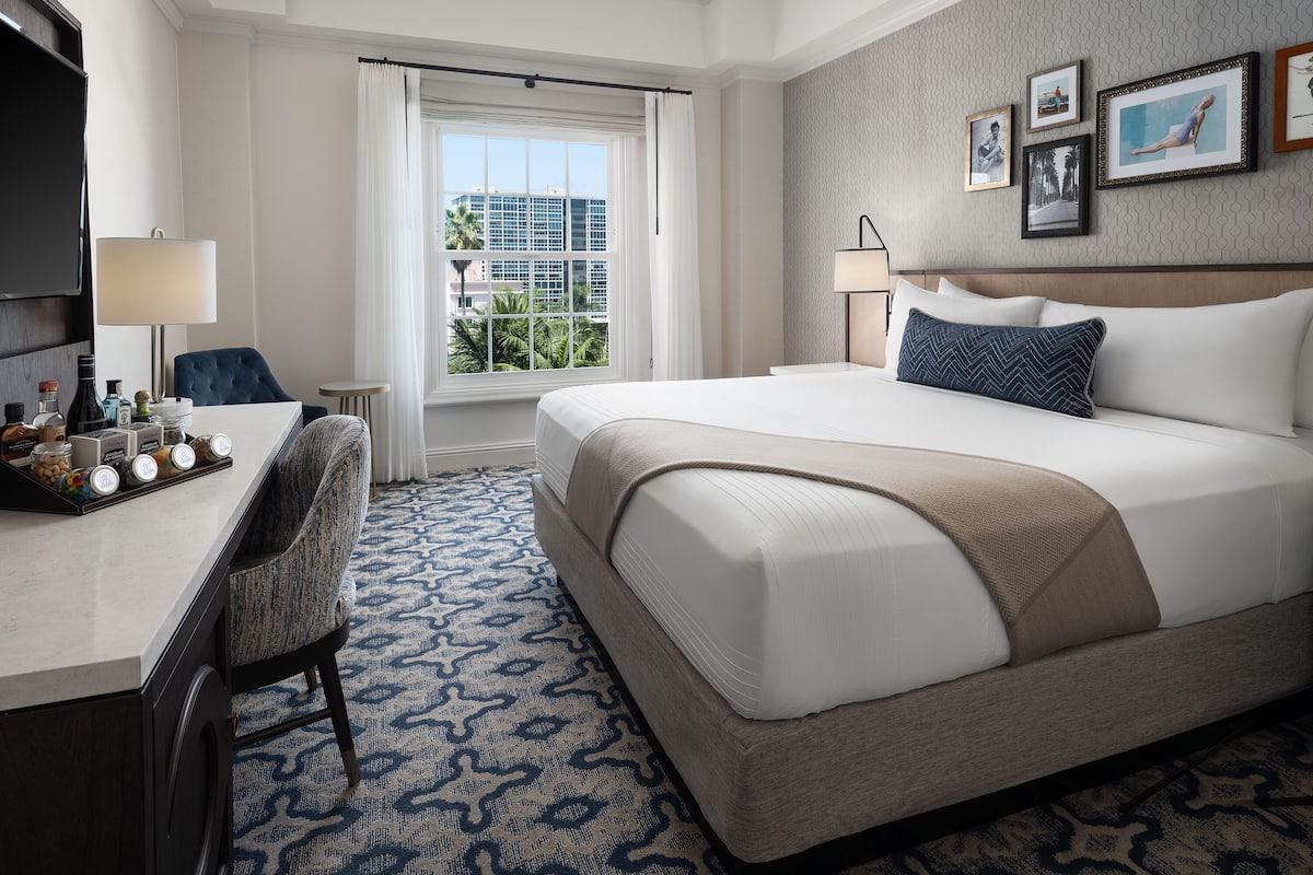 A hotel room with a large bed, a desk with a chair, a window with curtains, and framed pictures on the wall.
