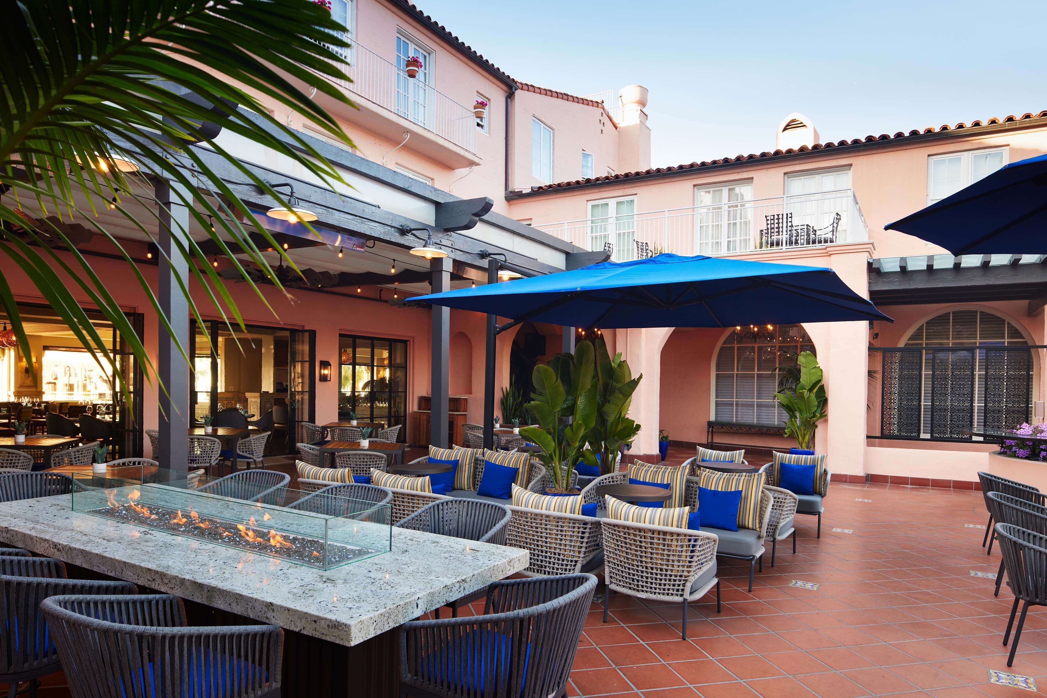 Eine Terrasse im Freien mit Korbmöbeln, einem Glastisch mit Feuerstelle und blauen Sonnenschirmen.