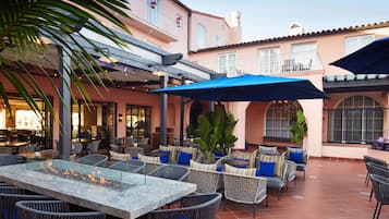 Eine Terrasse im Freien mit Korbmöbeln, einem Glastisch mit Feuerstelle und blauen Sonnenschirmen.