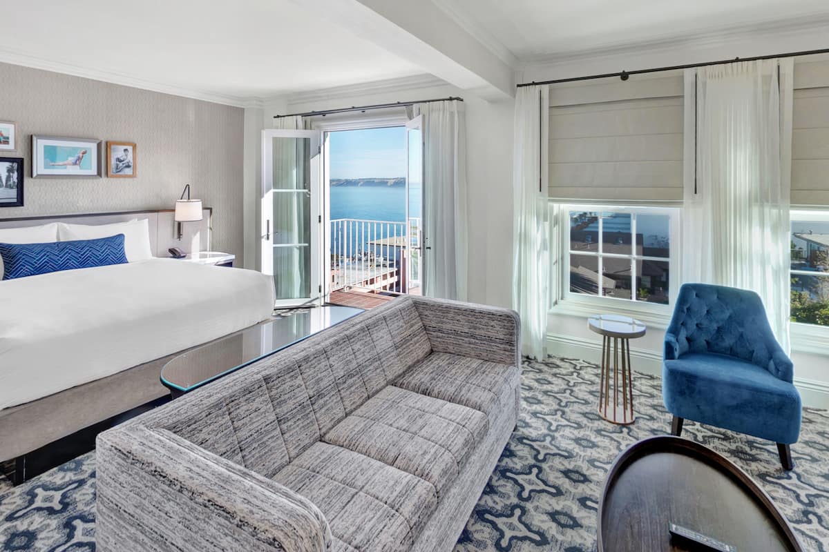 A hotel room with a large bed, a sofa, a chair, a small table, and a view of the sea through a balcony.