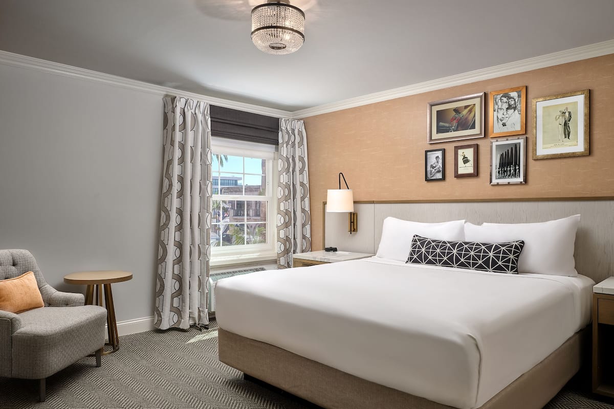 A hotel room with a large bed, a chair, a small table, and a window with curtains.