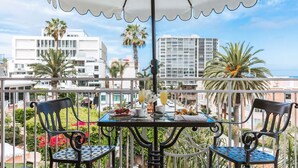 Varanda com duas cadeiras, uma mesa, guarda-sol branco e vista para a cidade, com edifícios e palmeiras.