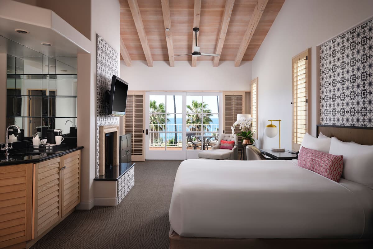 A hotel room with a large bed, a TV, a fireplace, a balcony with a view of the ocean, and a kitchen area.