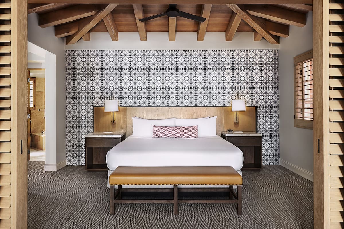 A bedroom with a bed, two bedside tables, a bench, a patterned wallpaper, and wooden beams on the ceiling.