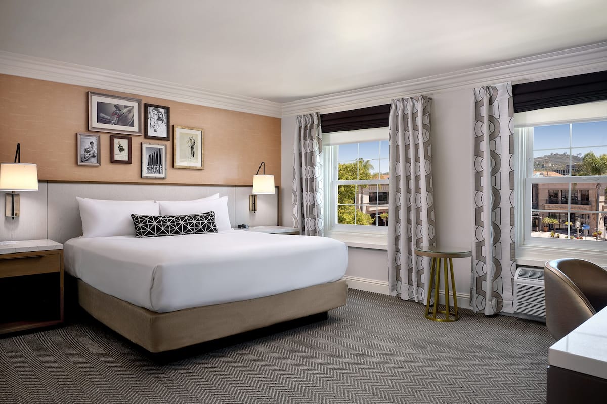 A hotel room with a large bed, two bedside tables, a desk, and a chair. There are framed pictures on the wall, a window with curtains, and a view of the outside.